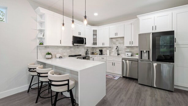 Cocina moderna con armarios blancos, una isla central con taburetes de bar, electrodomésticos de acero inoxidable e iluminación colgante.