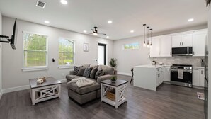 Una sala de estar moderna con un sofá gris, una mesa de centro blanca y una zona de cocina con armarios blancos y electrodomésticos de acero inoxidable.