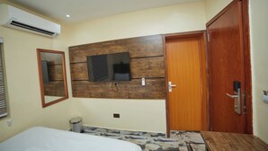 Una habitación de hotel con una cama, cabecera de madera, televisor montado en la pared, puerta de madera y aire acondicionado instalado.