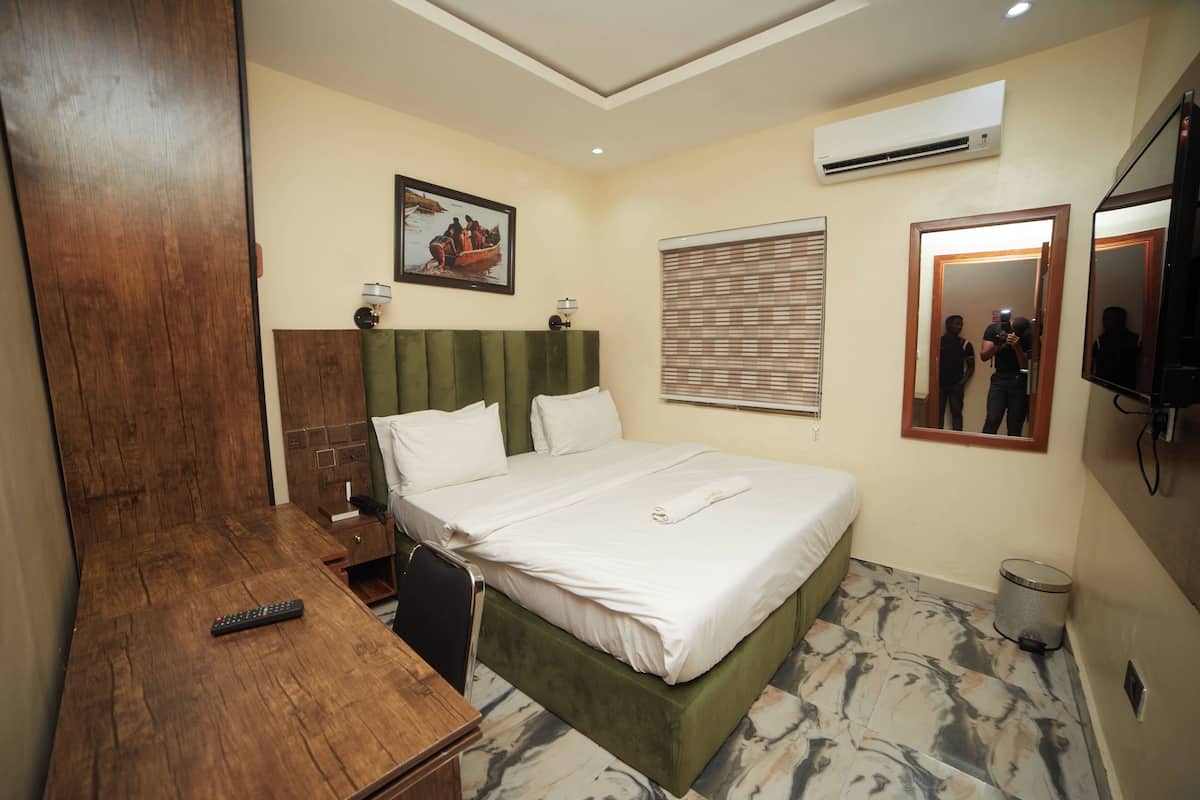 Une chambre d’hôtel comprenant un lit, un bureau en bois, une chaise, une table de chevet, un miroir, un climatiseur et une fenêtre avec des stores.