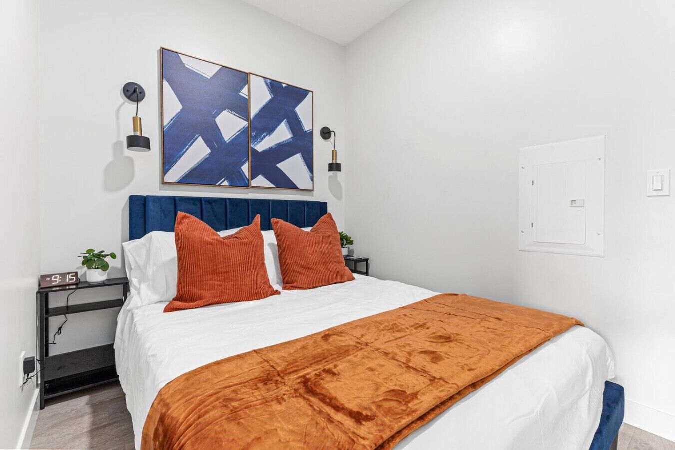 Une chambre à coucher avec un lit, une tête de lit bleue, deux lampes fixées au mur et un grand tableau abstrait.