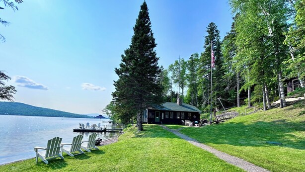 Una cabaña junto a un lago con un muelle, sillas y un sendero que la conecta.