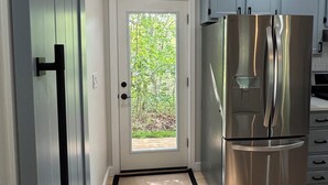 A modern kitchen with a stainless steel refrigerator, a sliding door, and a view through the door to a green outdoor area. - Spacious, Family-Friendly Edgartown Home – 3BR, Sleeps 14, Renovated in 2025 (Edgartown)
