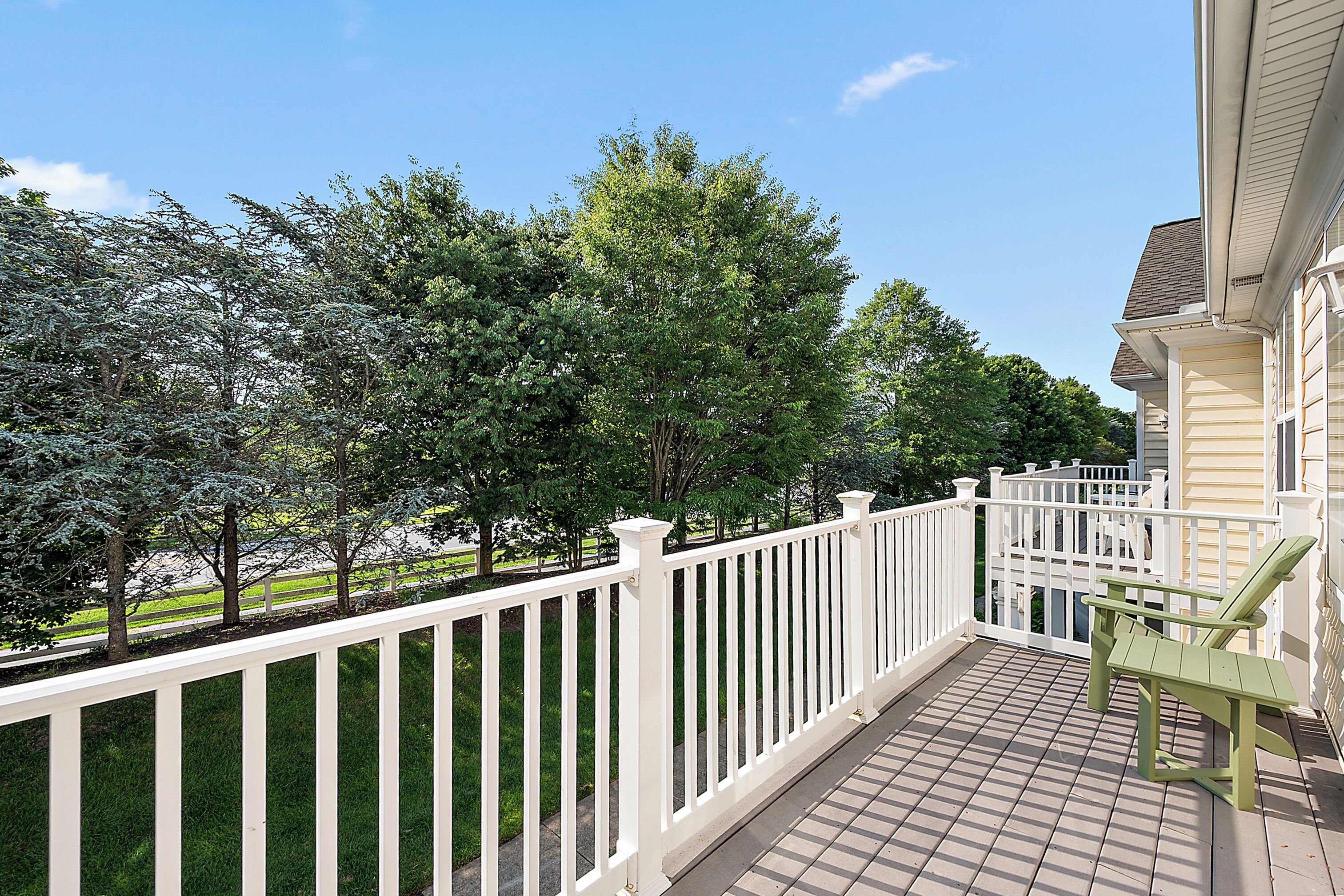 A wooden deck with a white railing, a green chair, and a view of trees and a road.