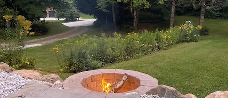 A fire pit surrounded by rocks and gravel, with a grassy area and trees in the background.