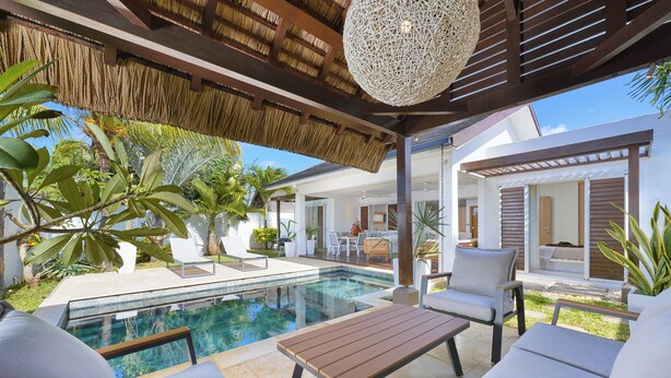 A poolside area with a wooden table and white lounge chairs, surrounded by tropical plants and a thatched roof structure.