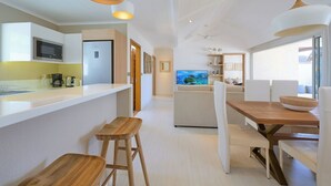 A modern kitchen with a microwave, refrigerator, and bar stools. There is a dining area with a wooden table and chairs, and a large window providing natural light.