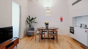 A modern living room with a wooden floor, a round dining table with chairs, a TV on a wooden stand, a grey armchair, a potted plant, and a pendant light.