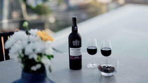 A bottle of Taylor's Very Old Single Harvest 1964 wine, two wine glasses, and a small bowl of chocolate on a table with a view of a cityscape in the background.