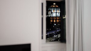 A room with a television on a wooden stand, a window with a view of a lit building at night, and a white door.