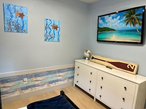 A bedroom with a bed, a dresser, two wall art pieces, a globe, and a TV displaying a beach scene.