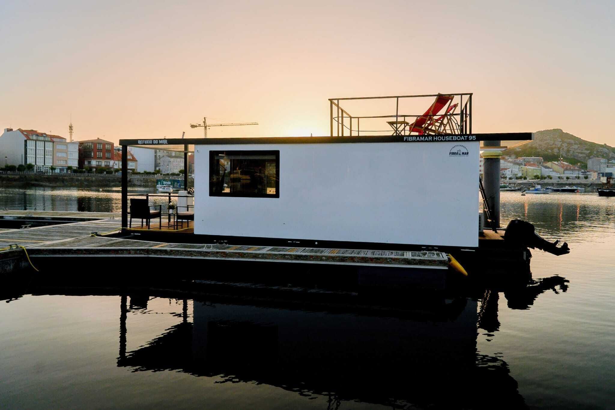 Una casa flotante amarrada en un puerto deportivo, con una pequeña terraza y una ventana.