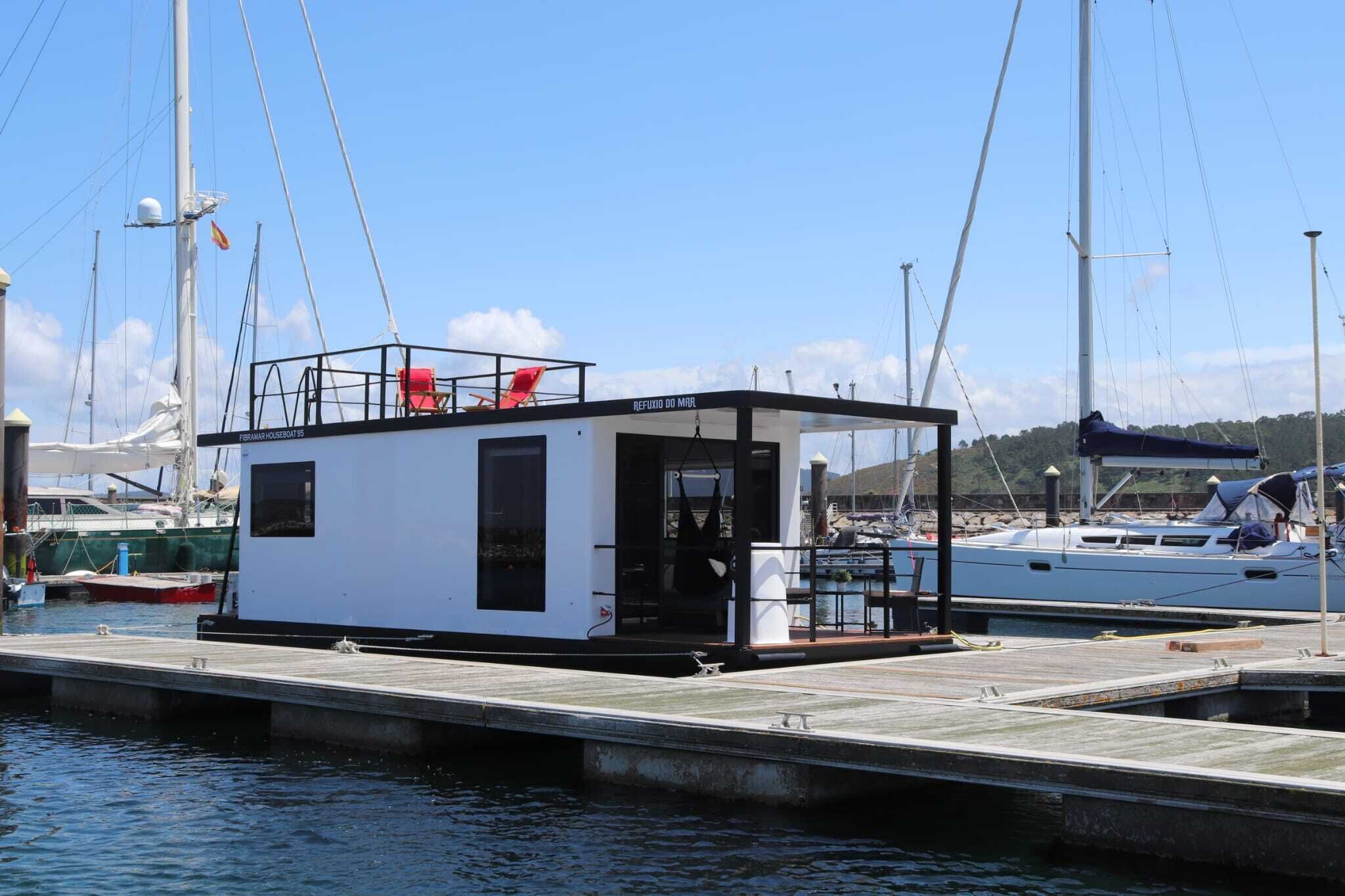 A small white building with a black roof, labeled "Refúgio do Mar," docked at a marina with other boats.
