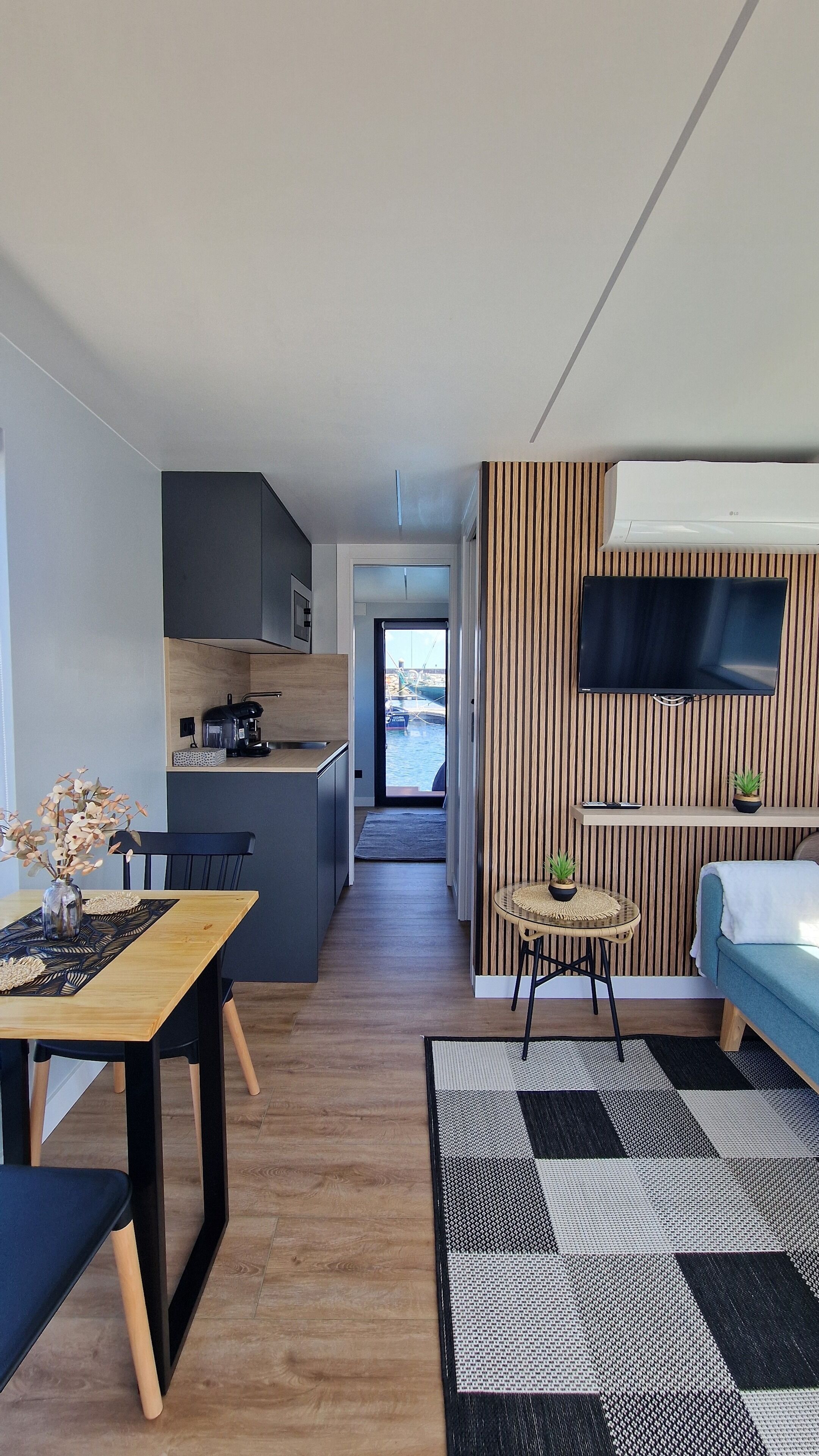 Une cuisine moderne équipée d’une machine à café, un coin repas avec une table et des chaises, et un salon avec un canapé et un téléviseur fixé au mur.
