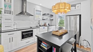 A modern kitchen with white cabinetry, a stainless steel range hood, a black countertop, and a hanging light fixture.