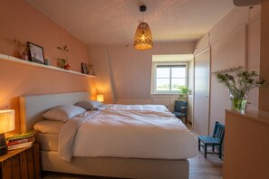 A bedroom with a bed, bedside lamps, a chair, a window, and a shelf with decorative items.