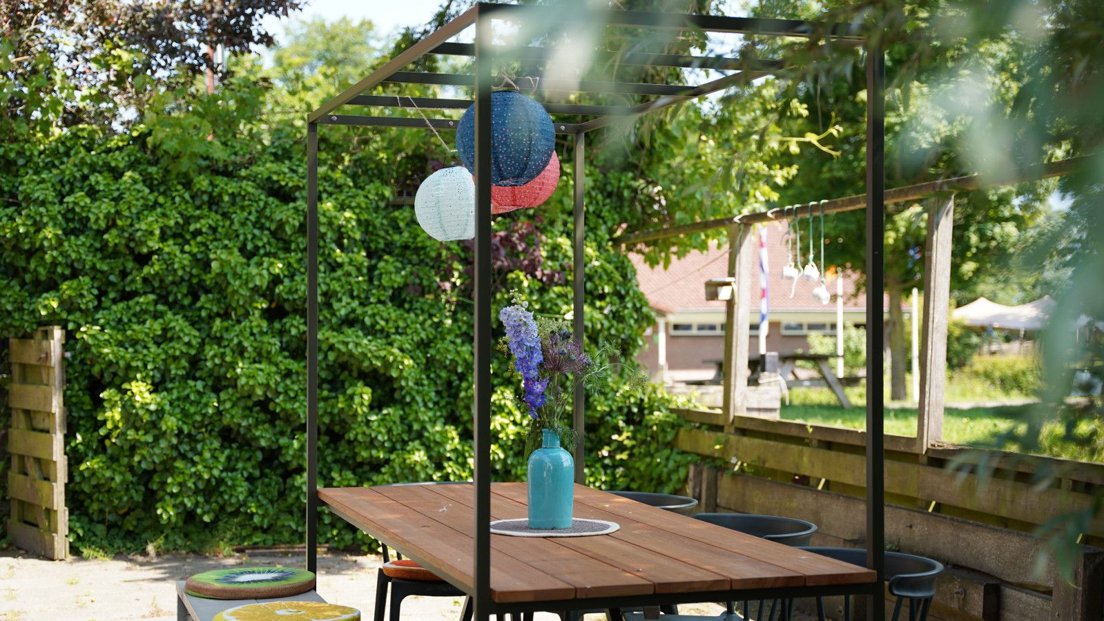En udendørs terrasse med et træbord og stole, en hængende lygte og en vase med blomster.