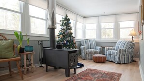 A cozy living room with a wood-burning stove, a Christmas tree, striped armchairs, and a large window.