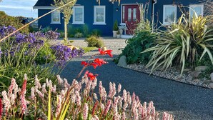 A house with a dark blue exterior, white trim, and a red door. There are various plants and flowers in the foreground, and a gravel driveway leading to the house. - Thistledown Lodge (Templetown)