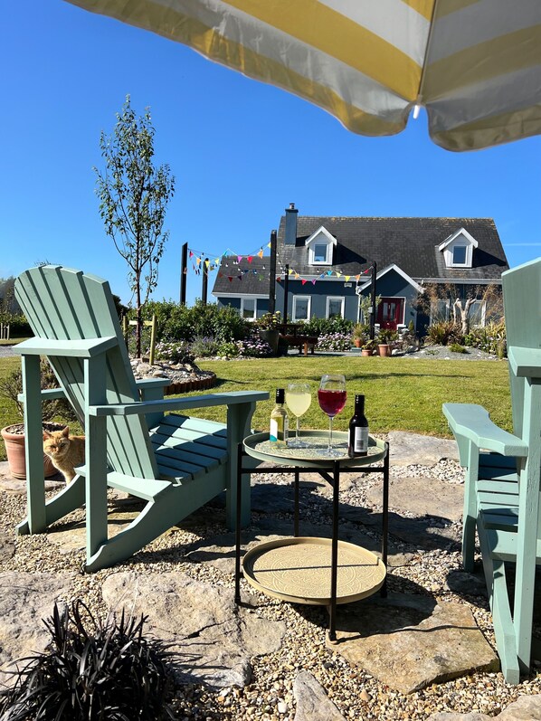 Un área de estar al aire libre con sillas Adirondack, una mesa con bebidas y una casa al fondo.