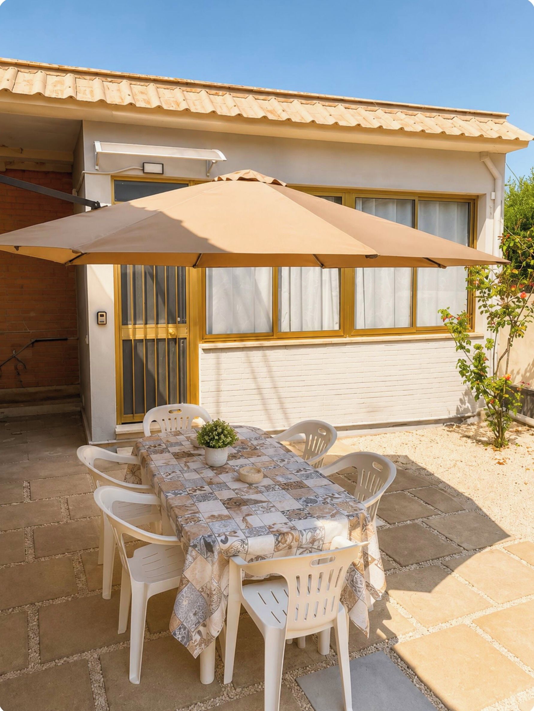 An outdoor patio with a table set for four, surrounded by white plastic chairs, a beige umbrella, and a potted plant.