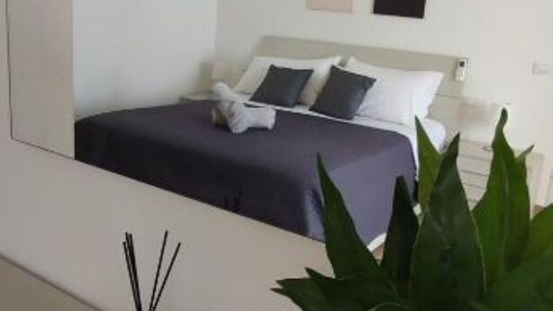 Una cama bien tendida con una colcha oscura y almohadas, una mesita de noche blanca y una planta en maceta al frente.