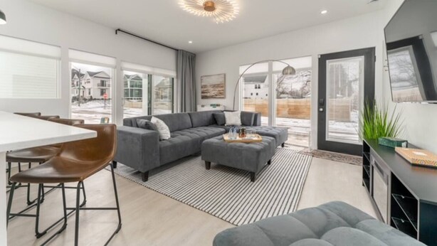 Un salon moderne avec un canapé d’angle, un coin repas comprenant une table blanche et des chaises marron, et une grande fenêtre donnant sur un paysage enneigé.