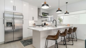 Une cuisine moderne avec des meubles blancs, un réfrigérateur en acier inoxydable, un îlot central doté de tabourets, des luminaires suspendus et une fenêtre avec des stores.