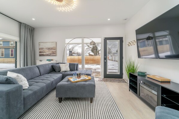A modern living room with a sectional sofa, a coffee table with drinks, a flat-screen TV, and a large window with a view of a wooden deck. - 3 Ensuite Suites · Mile to Lucas Oil (Indianapolis)