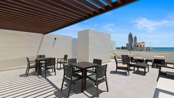 Un patio exterior con mesas y sillas, una pérgola y vistas al océano y edificios.