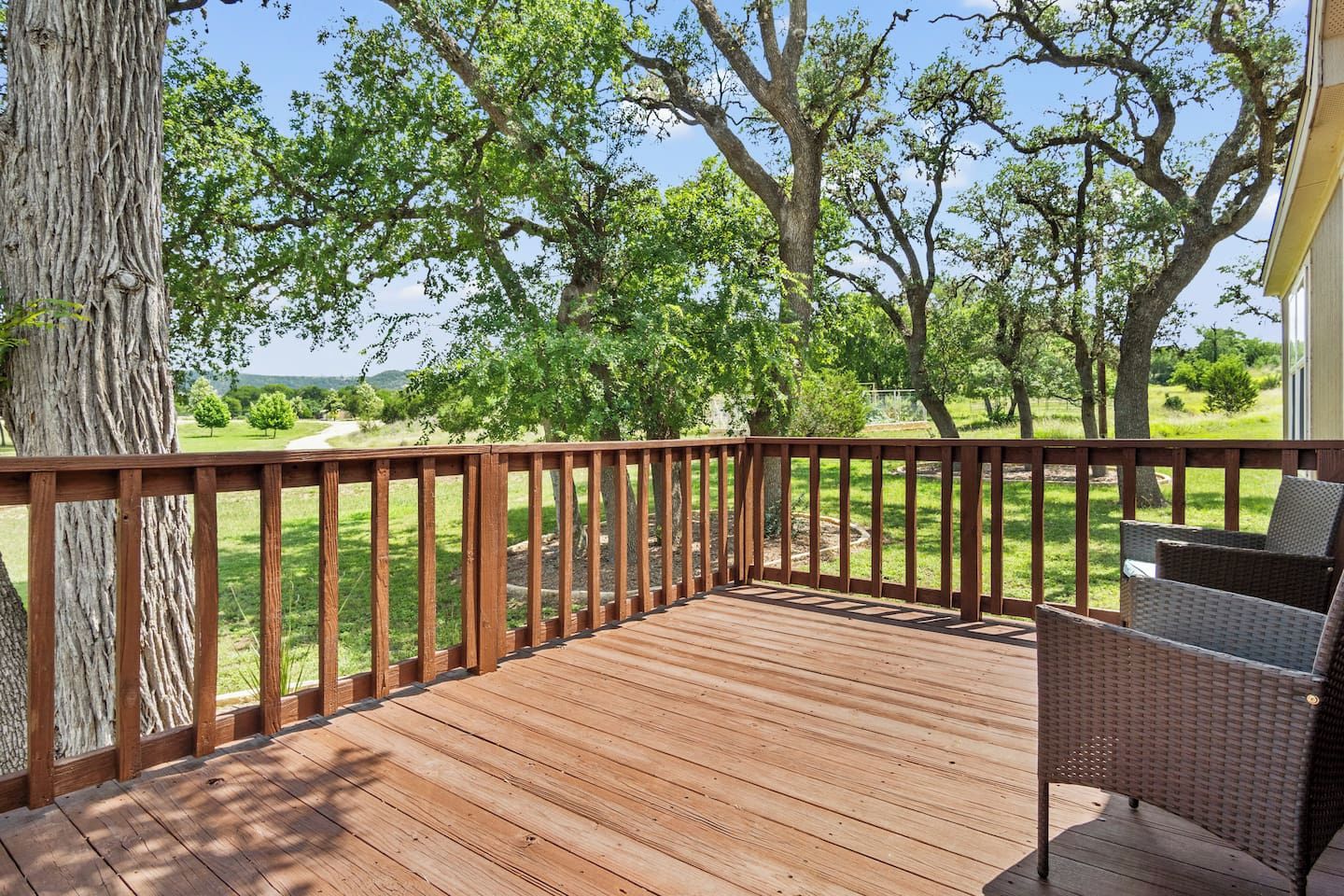 A wooden deck with a railing, wicker furniture, and a view of a grassy landscape with trees.