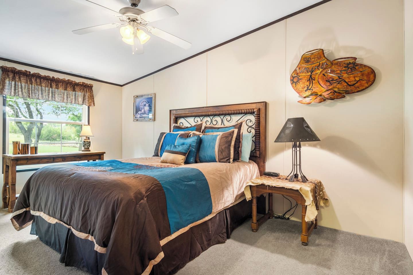 A bedroom with a bed, a nightstand, a lamp, a ceiling fan, a window with curtains, and a wall-mounted artwork.