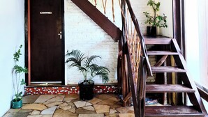 A staircase with a wooden handrail and rope accents, leading to a door with a black frame. There are potted plants on the floor and a white brick wall in the background. -  Pipa Universo (Tibau do Sul)