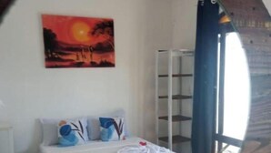A neatly made bed with white linens and blue pillows, a painting of a sunset on the wall, and a wooden-framed mirror reflecting the room. -  Pipa Universo (Tibau do Sul)