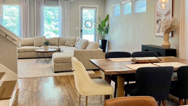 A modern living room with a sofa, dining table, and chairs. There are windows with curtains, a plant, and a ceiling fan. - Modern 3BR Townhome w/Garage - Walk to Midtown/Downtown Tallahassee & FSU (Tallahassee)