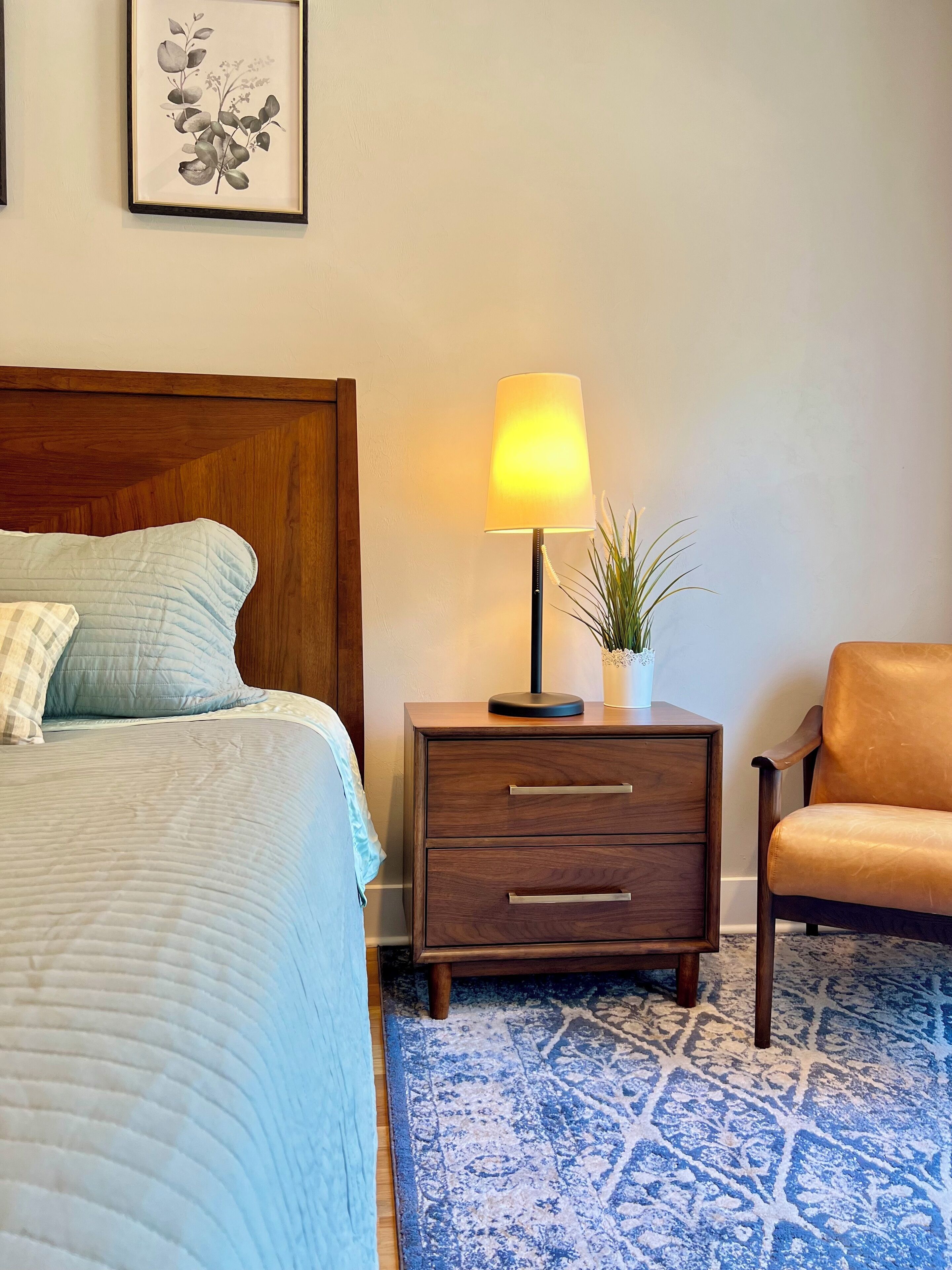 Una cama bien hecha con un edredón azul, una mesita de noche de madera con una lámpara, una pequeña planta en maceta y un sillón de cuero.