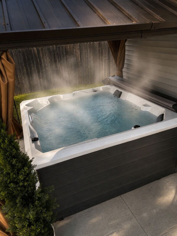 Un jacuzzi con terraza de madera y techo metálico, rodeado por una valla de madera y un árbol pequeño.