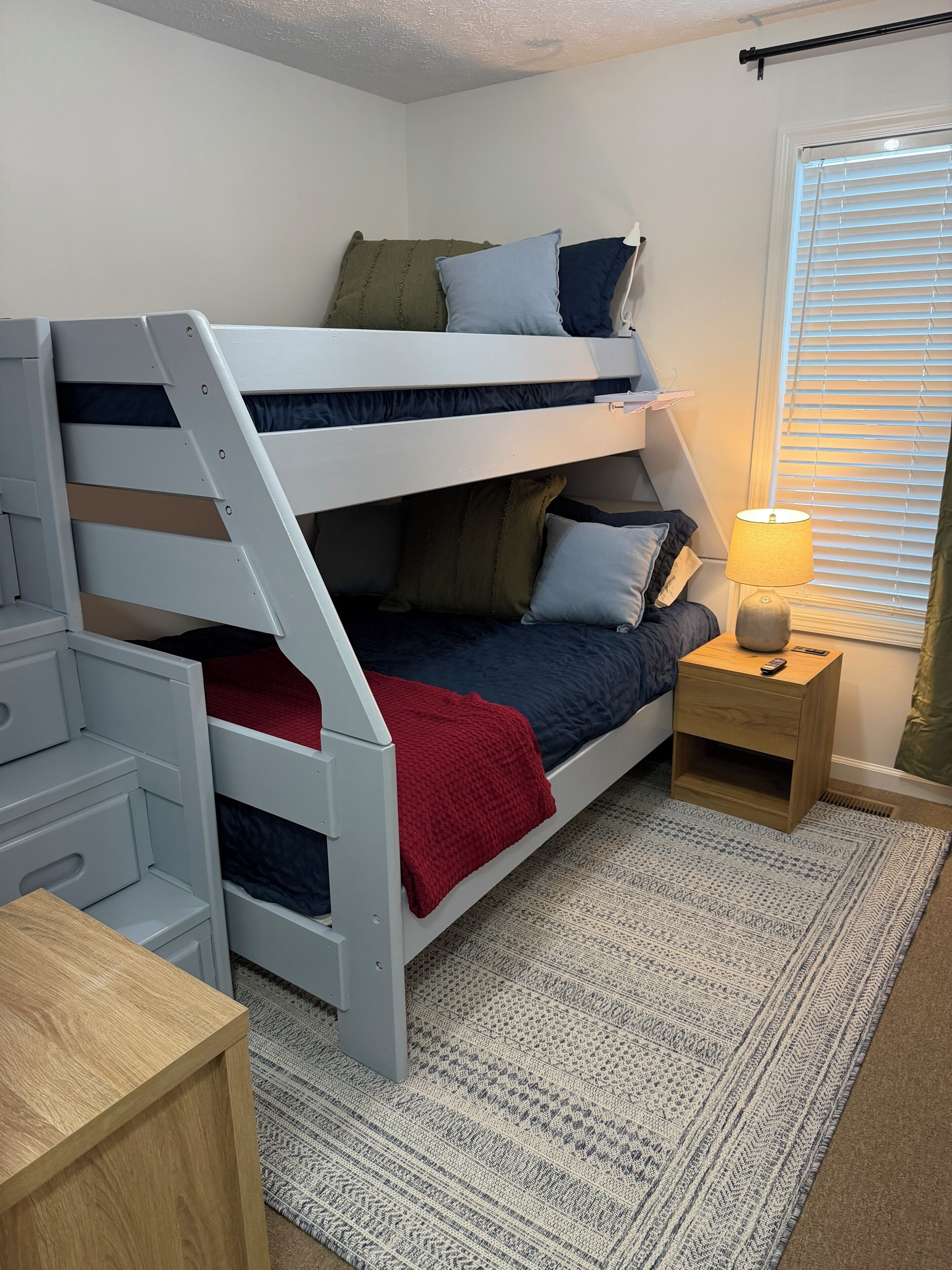 A bunk bed with a desk and drawers, a bedside table with a lamp, and a patterned rug.