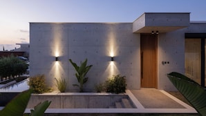 A modern building with a concrete facade, two illuminated wall lights, a wooden door, and a small garden area with plants and a concrete bench. - Designer Bungalow with Private Infinity Pool in Paphos (Chlorakas)