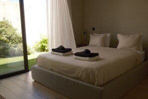 A large bed with white linens and two black towels on top, a bedside table with a lamp, and a view of greenery through a large window.