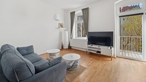 Una sala de estar moderna con un sofá gris, un televisor sobre un mueble blanco, una pequeña mesa redonda y un ventanal con cortinas.