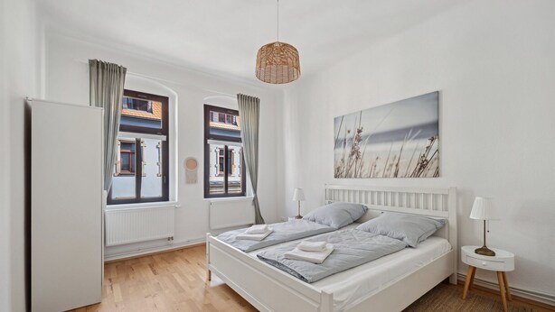 Un dormitorio con cama, mesita de noche, lámpara, ventana con cortinas y vista a edificios.