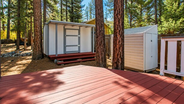 A deck with a wooden floor, surrounded by trees and two small sheds. - Cedar Pines (Big Bear City)
