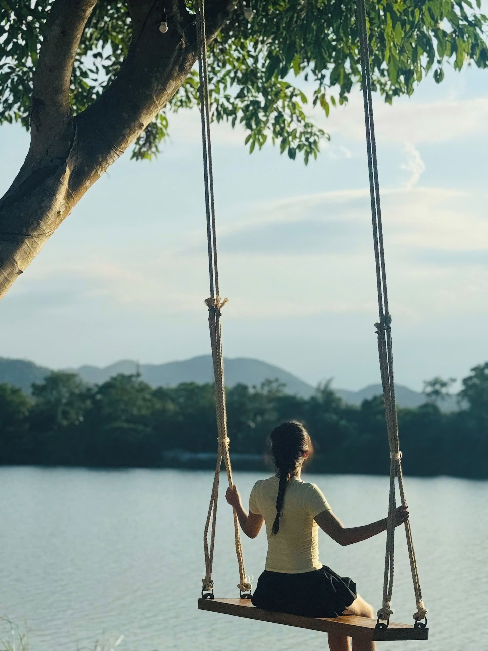 A person sitting on a swing suspended by ropes from a tree, overlooking a body of water and distant mountains.