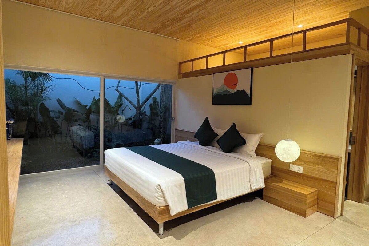 A modern bedroom with a large bed, a wooden headboard, a wall-mounted light, and a view of the outdoors.
