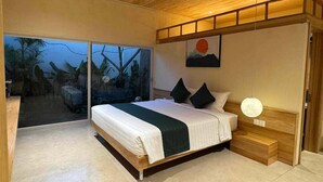 Ein modernes Schlafzimmer mit einem großen Bett, einer Schiebetür zu einem tropischen Ausblick und einem Sonnenuntergangsgemälde an der Wand.