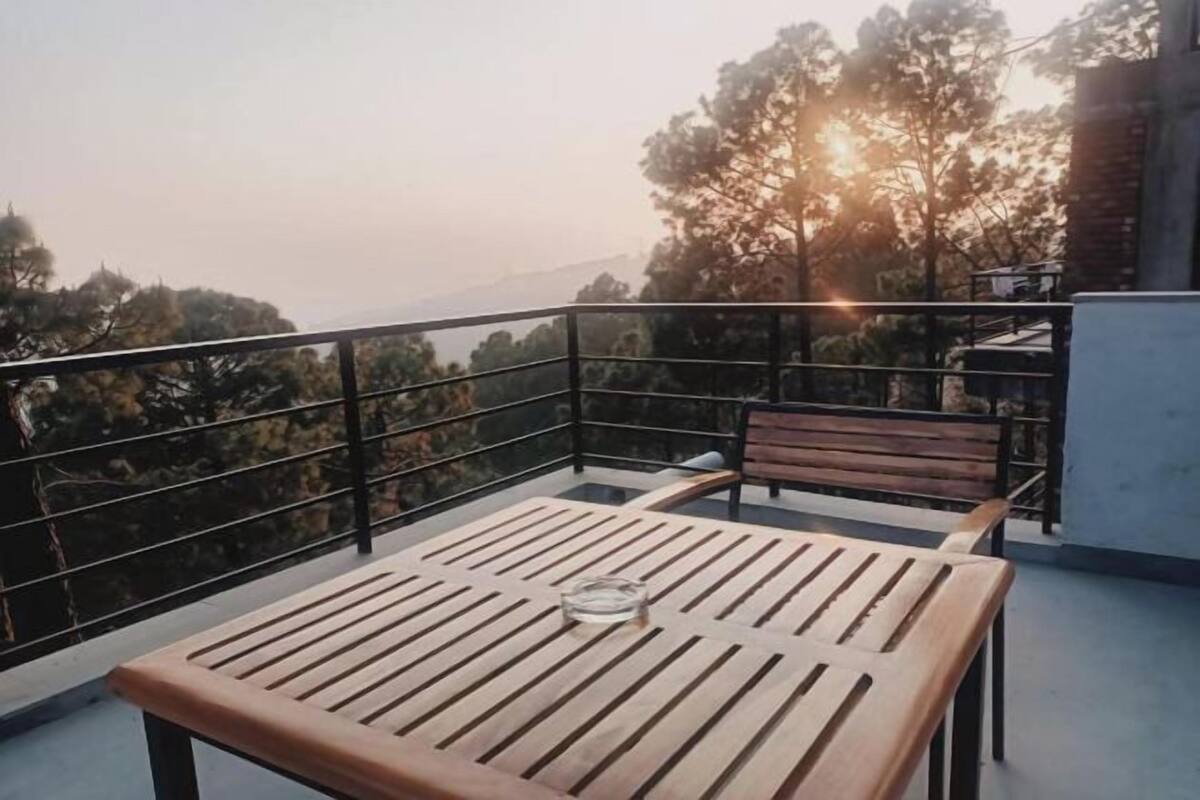 Un balcone con un tavolo e una panca di legno, un bicchiere d'acqua e vista su alberi e colline sullo sfondo.