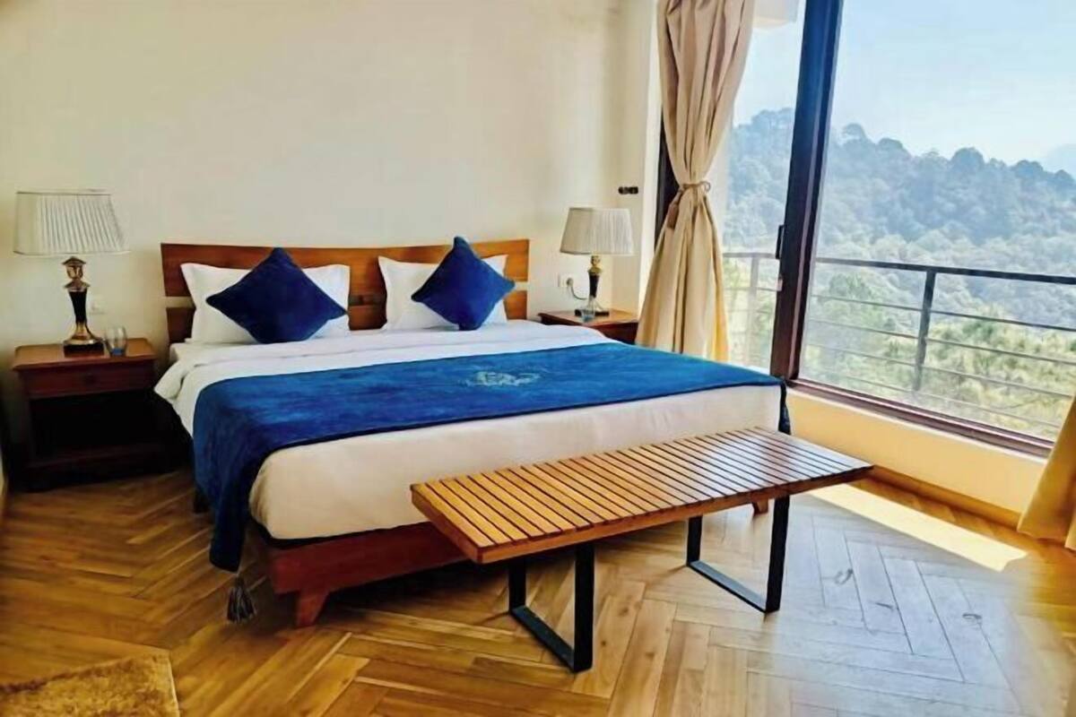 Una camera d'albergo con pavimento in legno, un letto con copriletto blu, due comodini con lampade, una panca in legno e un'ampia finestra con vista sulle colline.