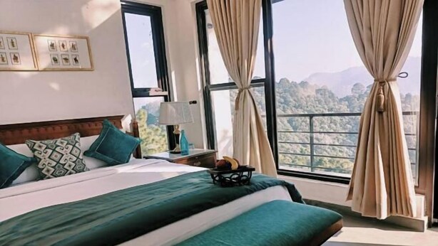 A hotel room with a large bed, a nightstand, a lamp, and a view of the mountains through the windows. - BluSalzz Escapade, Lansdowne - Uttarakhand (Lansdowne)