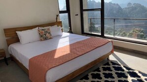 A bedroom with a large bed, a wooden headboard, and a view of mountains through large windows. - BluSalzz Escapade, Lansdowne - Uttarakhand (Lansdowne)